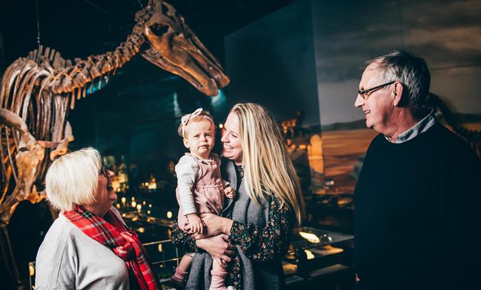 grandparents, mum and small girl. All smiling. Standing in front of a large dinosaur skeleton.